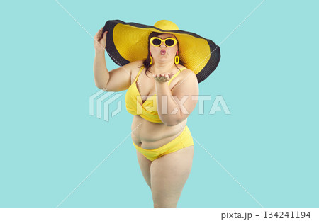 Fat woman in bikini, sunglasses and big beach hat giving air kiss on studio blue background. Fat woman in bikini, sunglasses and big beach hat giving air kiss on studio blue background. 134241194