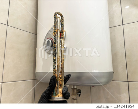 Plumber hand holding a boiler heating element covered with limescale, hard water residue, boiler maintenance and cleaning, scale removal, water heater repair 134241954