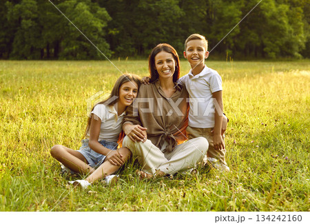 Happy mother with daughter and son sitting in the field on green grass and looking at camera. Happy mother with daughter and son sitting in the field on green grass and looking at camera. 134242160