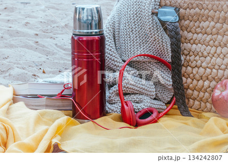 Picnic scene showing a red thermos, headphones, books, a knitted blanket, and a straw bag on a yellow fabric, enjoying outdoor leisure and relaxation Picnic scene showing a red thermos, headphones, books, a knitted blanket, and a straw bag on a yellow fabric, enjoying outdoor leisure and relaxation 134242807