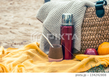 Relaxing outdoor picnic setup on the sand, featuring a thermos, mate gourd with bombilla, woven bag, and fresh fruits, enjoying leisure time Relaxing outdoor picnic setup on the sand, featuring a thermos, mate gourd with bombilla, woven bag, and fresh fruits, enjoying leisure time 134242808