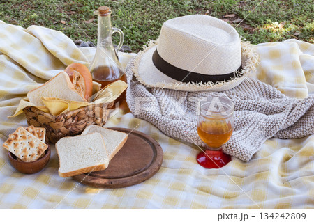 Checkered picnic blanket on green grass with straw hat, bottle and glass of amber drink, crackers and assorted breads in a basket for a relaxing summer outdoor snack 134242809