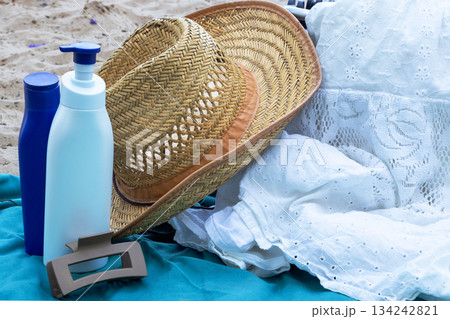 Summer beach essentials including a straw hat. Sunscreen bottles offering sun protection. A hair claw. And a white lace dress laying on a turquoise towel on sand 134242821
