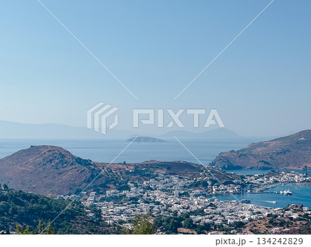 Greek island town of whitewashed buildings cascading to a busy harbor, boats bobbing in turquoise aegean waters beneath sunlit hills and distant mountains perfect travel escape 134242829