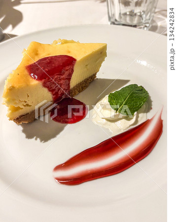 Cheesecake slice with creamy texture and graham cracker crust, topped with red berry sauce, whipped cream and mint on a white plate elegant dessert indulgence 134242834