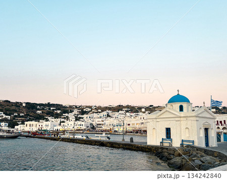 Mykonos island experiencing the sunset glow, presenting a whitewashed orthodox church with a blue dome by the harbor and traditional cycladic architecture along the coast Mykonos island experiencing the sunset glow, presenting a whitewashed orthodox church with a blue dome by the harbor and traditional cycladic architecture along the coast 134242840