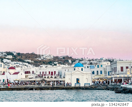 Iconic Mykonos island at sunset , presenting a whitewashed orthodox church with a blue dome by the harbor and traditional cycladic architecture 134242847