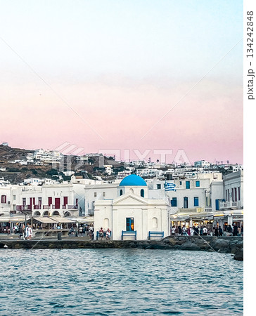 Verrical shot of Mykonos island at sunset , presenting a whitewashed orthodox church with a blue dome and traditional cycladic architecture 134242848