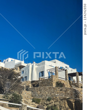 Mykonos island architecture featuring traditional cycladic whitewashed buildings with blue shutters and stone walls climbing a rugged hillside under a vibrant clear sky 134242850