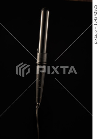 A hair curling iron is shown with a focus on its sleek design. The curling iron is turned off and positioned against a dark surface ready for use in creating waves or curls. 134242925