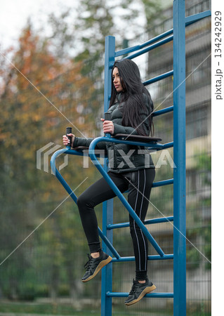 brunette woman is working out at a stadium. She is using a pull up bar while dressed in black workout clothes. Trees are in the background showing the season. brunette woman is working out at a stadium. She is using a pull up bar while dressed in black workout clothes. Trees are in the background showing the season. 134242929