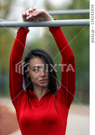 Brunette woman is at a stadium holding onto a bar with her arms raised. It is daytime and she appears focused on her workout. The atmosphere shows outdoor activity. Brunette woman is at a stadium holding onto a bar with her arms raised. It is daytime and she appears focused on her workout. The atmosphere shows outdoor activity. 134242939