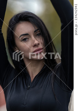brunette woman stands at the stadium raising her arms in a playful manner. She is outdoors enjoying a sunny day surrounded by stadium views and lively energy. 134242941