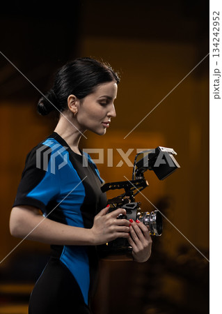 brunette woman stands in a studio holding a camera. She appears focused on her task. The lighting enhances her profile as she prepares to capture images. 134242952