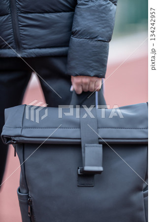 woman with brown hair holds a black backpack while walking on a path. She wears a black jacket and appears focused on her direction as she walks. 134242957