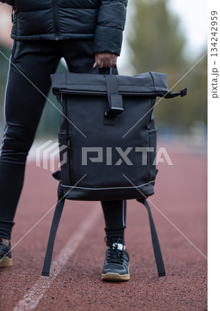 brunette wears a black jacket and holds a black backpack. She stands on a running track during early morning. The surrounding area shows trees and sports equipment. brunette wears a black jacket and holds a black backpack. She stands on a running track during early morning. The surrounding area shows trees and sports equipment. 134242959