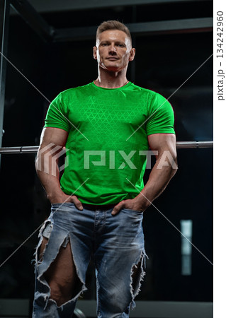 man stands near a Smith machine in a gym wearing a green shirt and ripped jeans. He has a relaxed expression as he prepares for his workout early in the morning. 134242960