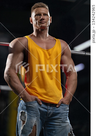 man in a gym stands near a Smith machine. He has a focused expression and wears a tank top and ripped jeans. The setting shows weights and gym equipment around him. 134242965
