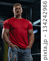 man with a muscular build stands near a Smith machine in a gym. He is wearing a red shirt and has his hands in his pockets. The lighting highlights his features. 134242966