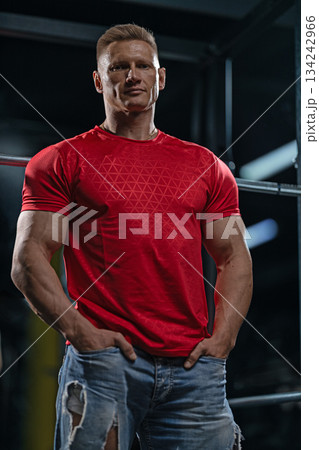 man with a muscular build stands near a Smith machine in a gym. He is wearing a red shirt and has his hands in his pockets. The lighting highlights his features. 134242966