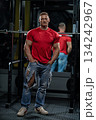 man stands by a Smith machine in a gym. He wears a red shirt and ripped jeans. He appears focused and ready for the next exercise. Gym equipment is visible in the background. 134242967