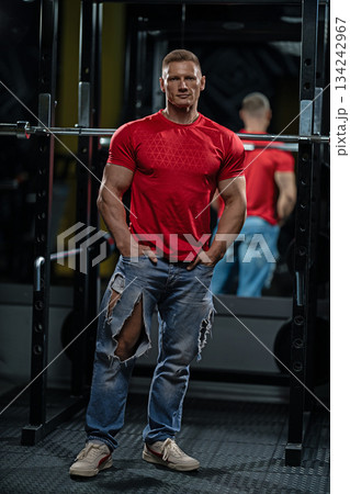 man stands by a Smith machine in a gym. He wears a red shirt and ripped jeans. He appears focused and ready for the next exercise. Gym equipment is visible in the background. 134242967