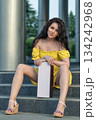 brunette woman in a yellow dress is sitting on the steps. She holds a box while enjoying the sunlight. The setting features modern architecture and greenery nearby. 134242968