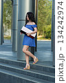 brunette woman in a dress walks down steps while carrying a box. The setting is an outdoor area with modern architecture and greenery. It is daytime. 134242974