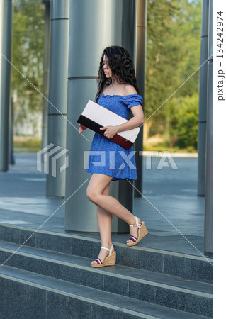 brunette woman in a dress walks down steps while carrying a box. The setting is an outdoor area with modern architecture and greenery. It is daytime. 134242974