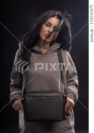 brunette woman is wearing a hoodie and holding a bag while posing in a dimly lit studio. Her hair is flowing around her as she looks at the camera. 134242975