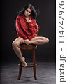 brunette woman wearing a hoodie is sitting on a wooden chair with her legs stretched out. She is posing with her hands creating an artistic look in a studio setting. 134242976