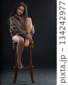 brunette woman sits with one leg curled under her on a chair. She wears a hoodie and looks directly at the viewer in a dark room with soft lighting. Her expression is relaxed. 134242977