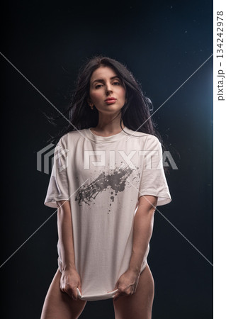 brunette woman stands in a studio with soft light. She holds her white T shirt with both hands looking directly at the camera. The setting is dim and focused on her. 134242978
