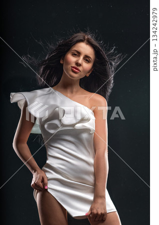 A brunette woman stands confidently wearing a short white dress. Her hair flows freely as she poses against a dark backdrop highlighting her fashion choice and personality. 134242979