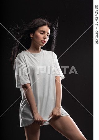 brunette stands in a white T shirt against a dark background. She looks thoughtful while holding the hem of her shirt. The studio light highlights her features and creates a dramatic atmosphere. 134242980