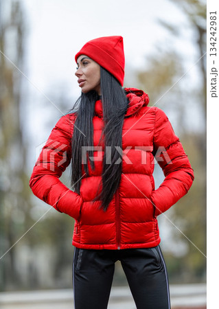brunette woman stands at the stadium with a red jacket and matching hat. She has her hands in her pockets and looks confidently ahead. The atmosphere is cloudy and cool. 134242981