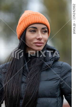 brunette woman stands at a stadium wearing a black jacket and an orange hat. She looks around while attending an outdoor event with many people and activities happening. 134242982