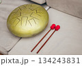 Golden handpan drum with geometric sound holes rests on a sofa, by two vibrant red mallets 134243831
