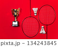 Golden Trophy Stands Beside Two Badminton Rackets And Two White Shuttlecocks Against Red Background 134243845