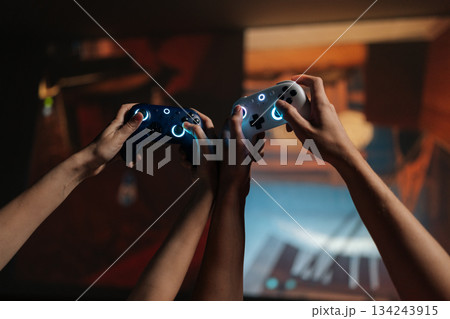 Young couple hands holding illuminated wireless game controllers, actively competing in online video game tournament, enjoying leisure time and modern digital entertainment together at home. Young couple hands holding illuminated wireless game controllers, actively competing in online video game tournament, enjoying leisure time and modern digital entertainment together at home. 134243915