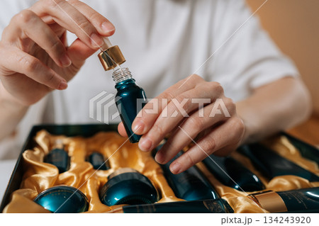Woman hands carefully holding dropper with liquid serum above a small bottle, part of a luxurious cosmetic gift set nestled in golden fabric, emphasizing beauty rituals and personal care. Woman hands carefully holding dropper with liquid serum above a small bottle, part of a luxurious cosmetic gift set nestled in golden fabric, emphasizing beauty rituals and personal care. 134243920