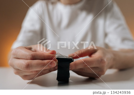 Close-up of female hands removing clear protective film from screen of smartwatch with fabric strap sitting at white table. Concept of device protection, repair, caring for personal technology. 134243936