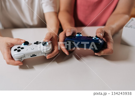 Close-up of happy relaxed couple holding using white and blue wireless controllers, engaging in gaming session sitting at white table, enjoying modern entertainment and companionship. 134243938