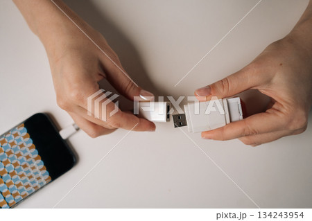 Top view of young adult woman hands connecting USB flash drive to smartphone using adapter cable sitting at white table, transferring data or charging device, close-up. Concept of modern technology. 134243954