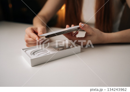 Cropped shot of woman unboxing modern smartphone, revealing sleek device on minimalist white table, displaying anticipation during initial technology reveal. Concept of cellphone unpacking process 134243962