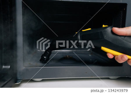Close-up of person cleaning microwave oven interior with steam cleaner appliance for hygiene and disinfection, removing grease and food residue from appliance surface. Concept of kitchen hygiene. Close-up of person cleaning microwave oven interior with steam cleaner appliance for hygiene and disinfection, removing grease and food residue from appliance surface. Concept of kitchen hygiene. 134243979