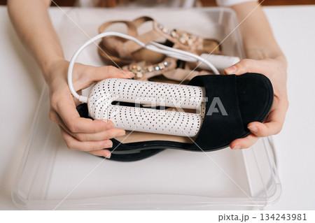 Top view of unrecognizable woman using domestic electric shoe dryer device, drying black footwear in plastic container, caring for suede shoes and sandals to remove moisture and unpleasant odor home. 134243981