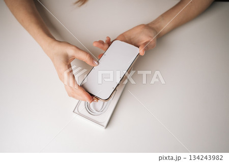 Top view of female hands unpacking new mobile phone from box on white table, eagerly holding device and preparing for exciting first-time setup experience. Concept of smartphone unboxing process 134243982