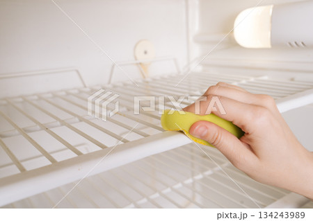 Close-up of unrecognizable female hand wiping down interior shelves of empty refrigerator with yellow sponge, performing thorough cleaning to ensure hygiene and maintain cleanliness in kitchen space. 134243989
