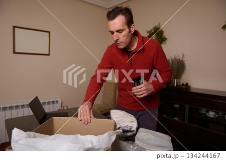 Man Packing Box At Home With Clothes In Living Room, Preparing Shipment Man Packing Box At Home With Clothes In Living Room, Preparing Shipment 134244167